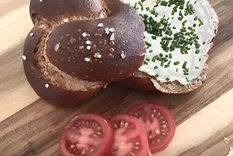 Vollkornbrezeln und -laugengebäck Ein Brötchen mit Frischkäse und Schnittlauch, serviert mit Tomatenscheiben.