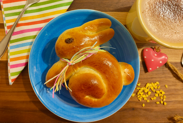 Osterhäschen und Mohnschnecken Hefeosterhäschen mit buntem Band, serviert auf einem blauen Teller, neben einer Tasse.