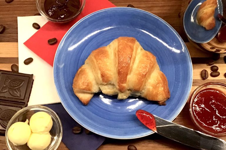 Croissants und Pains au Chocolat Ein Croissant auf einem blauen Teller, umgeben von Marmelade, Schokolade und Butterkugeln.
